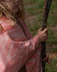 Kleinkind hat personalisierte lilimoon Babydecke gestrickt aus Baumwolle und Kaschmir in Korallenrosa und Cremeweiss wärmend um die Schultern gelegt und spielt mit einem Stock in der Natur.