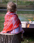 Kleinkind sitzt mit einer personalisierten lilimoon Babydecke gestrickt aus Baumwolle und Kaschmir in Rot und Flieder auf einem Baumstumpf vor einer Feuerstelle in der Natur.