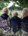 Drei Kleinkinder sitzen in der Natur auf einem Baumstamm und tragen gestrickte lilimoon Babydecken mit Sternen und Blumen wärmend über ihren Schultern.