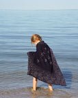 Kleinkind steht barfuss im Wattmeer der Nordsee und hat eine gestrickte lilimoon Babydecke mit Sternen und dem Sternzeichen Löwe als Umhang über die Schultern gelegt.