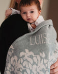 Ein Baby wird von seiner Mama auf dem Arm getragen umhüllt von einer wärmenden und personalisierten lilimoon Babydecke in hellgrau und cremeweiss mit Blütenmuster und eingestricktem Namen des Kindes.