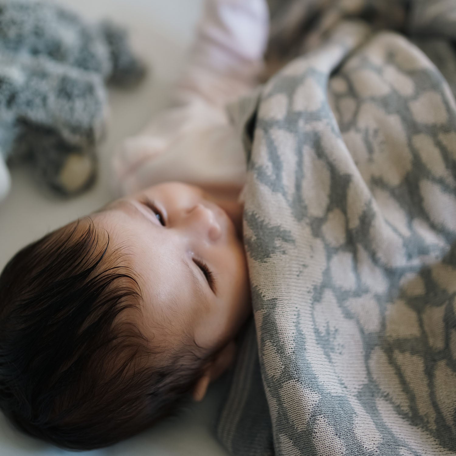Ein Baby liegt zufrieden und zugedeckt mit einer kuscheligen lilimoon Babydecke in hellgrau und cremeweiss mit Blütenmuster in seinem Babybett.