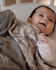 Ein fröhliches Baby liegt zugedeckt mit einer personalisierten lilimoon Babydecke in hellgrau und cremeweiss mit Blütenmuster im Kinderbett.