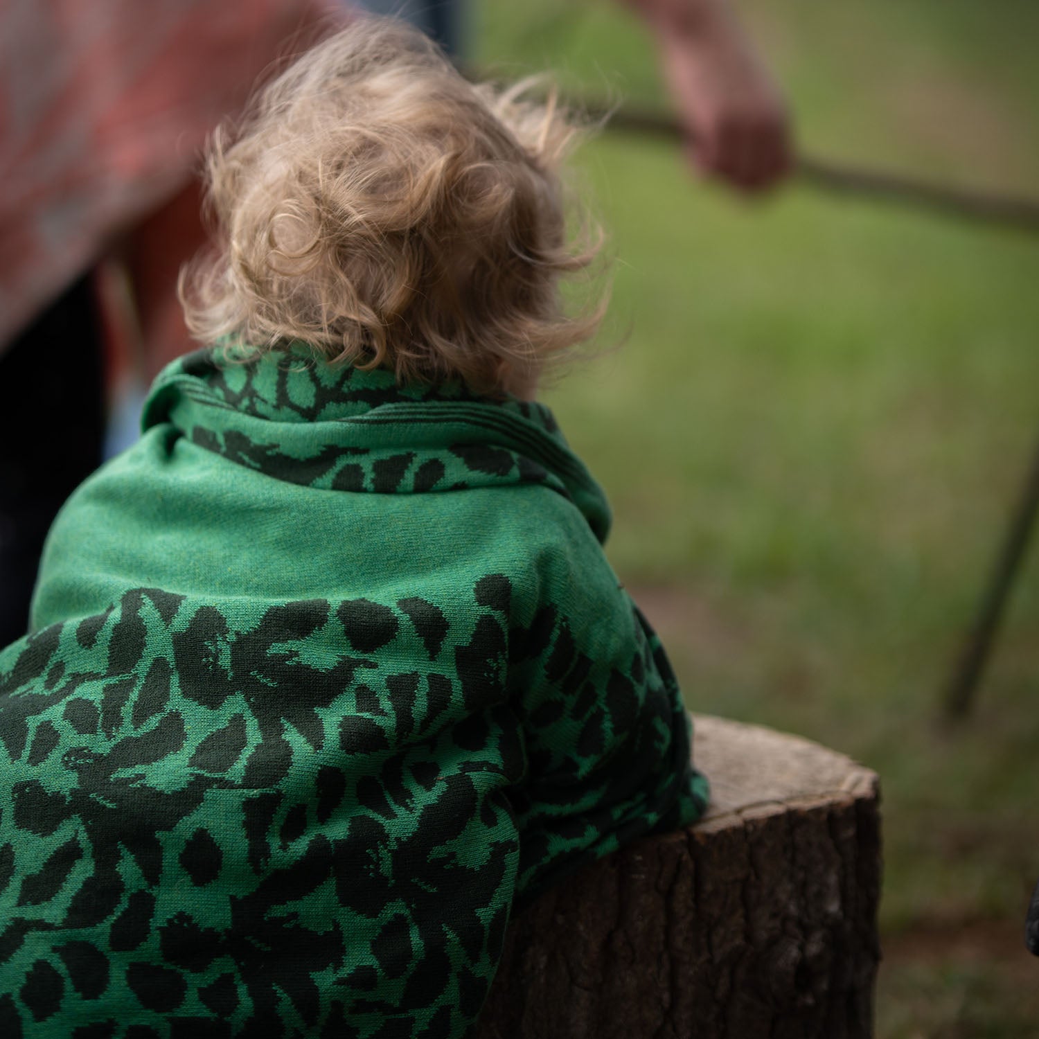 Ein Kleinkind stützt sich in der Natur auf einen Baumstumpf und hat sich eine grüne lilimoon Babydecke mit Blütenmuster wärmend um die Schultern gelegt.
