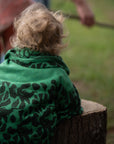 Ein Kleinkind stützt sich in der Natur auf einen Baumstumpf und hat sich eine grüne lilimoon Babydecke mit Blütenmuster wärmend um die Schultern gelegt.