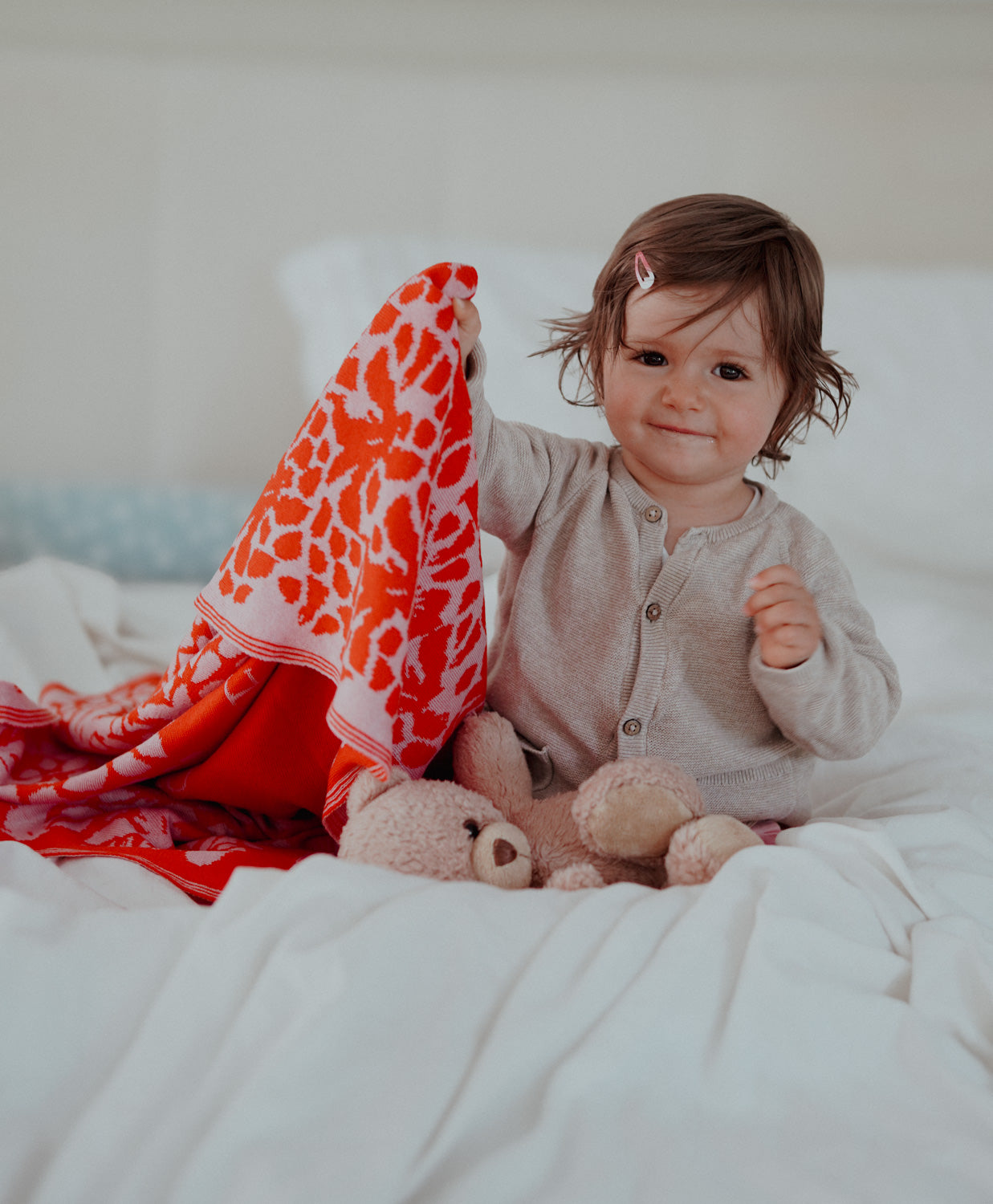 Ein kleines Mädchen sitzt fröhlich auf einem Bett und zeigt stolz ihre gestrickte lilimoon Babydecke in leuchtendem rot und rosa mit Blütenmuster.