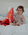 Ein kleines Mädchen sitzt fröhlich auf einem Bett und zeigt stolz ihre gestrickte lilimoon Babydecke in leuchtendem rot und rosa mit Blütenmuster.