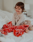 Ein fröhliches Kleinkind sitzt auf dem Bett und ist zugedeckt mit einer leuchtend rot und rosa farben lilimoon babydecke mit blütenmuster.