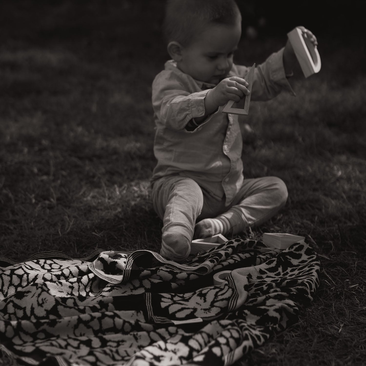 Ein kleiner Junge sitzt im Garten auf einer lilimoon Babydecke in schwarz und weiß mit Blütenmuster und spielt  mit Holzspielzeug.