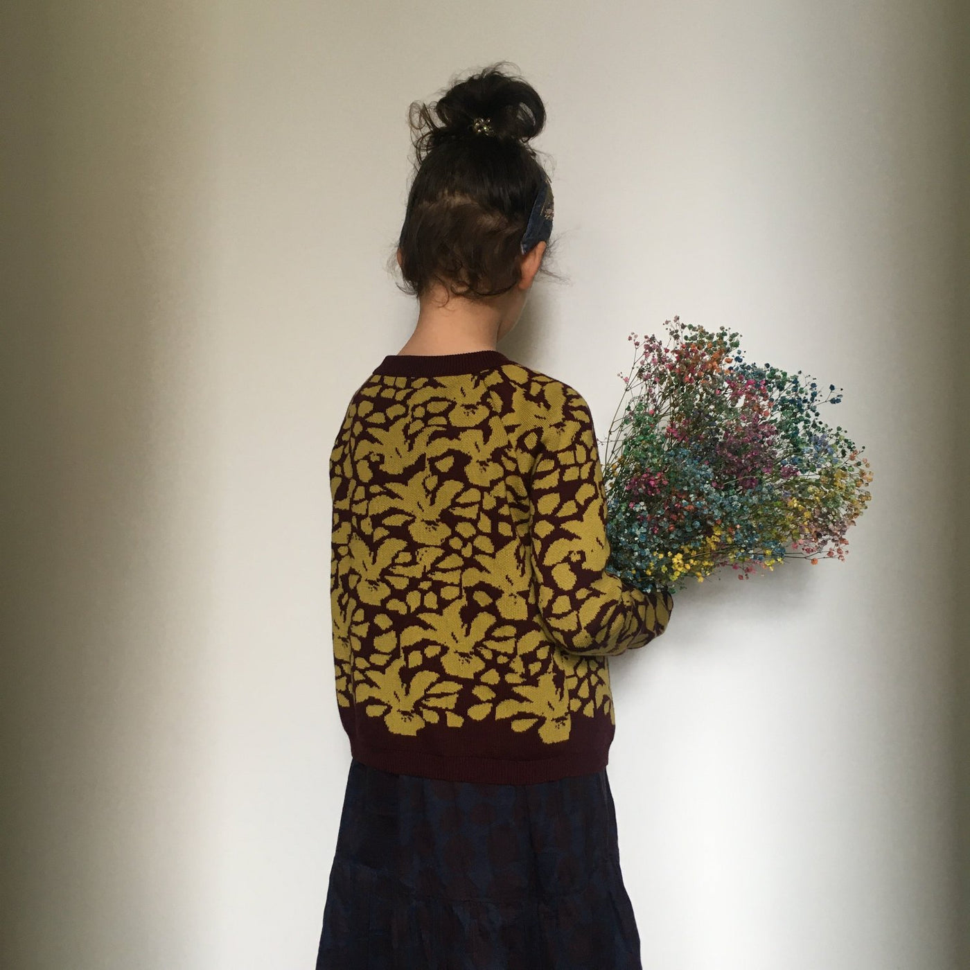 Junges Mädchen trägt eine gestrickte Jacke in Senfgelb und Burgund und hält einen Strauß Trockenblumen im Arm