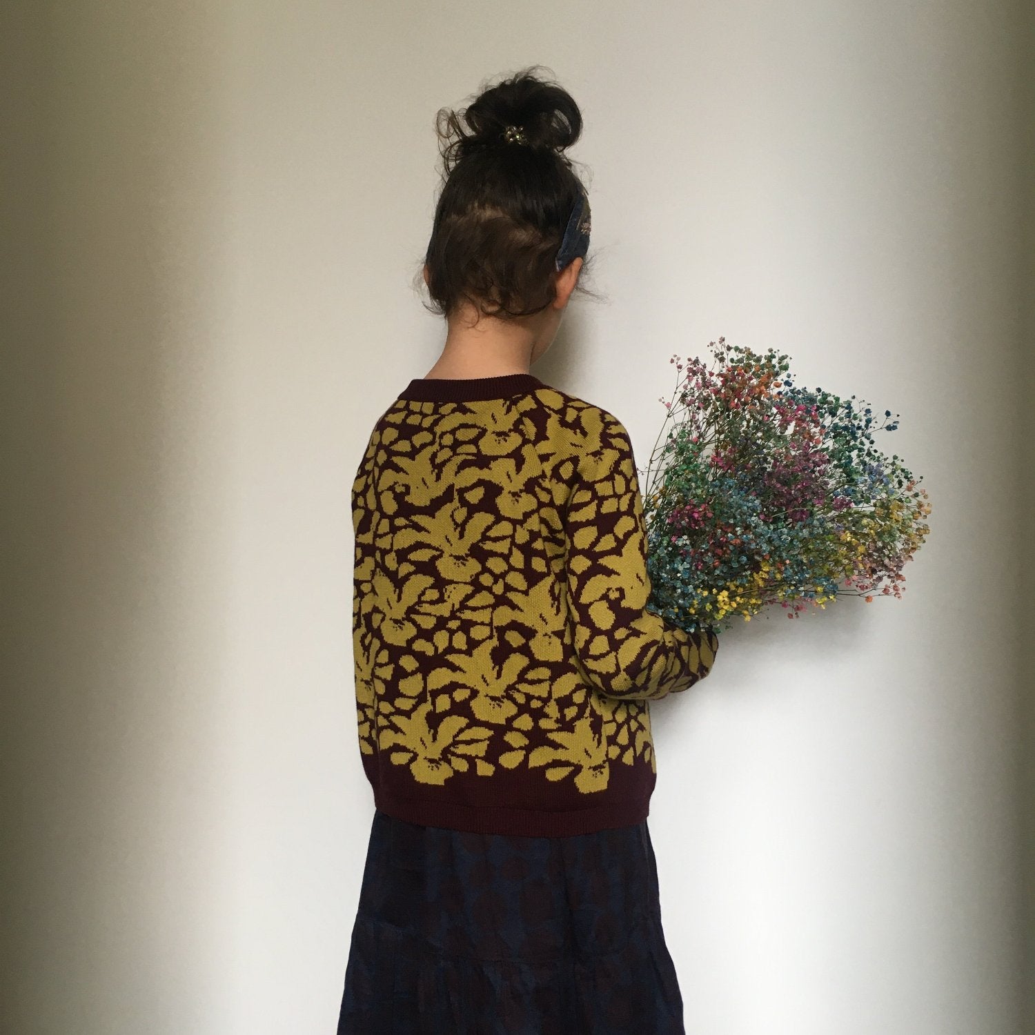 Junges Mädchen trägt eine gestrickte Jacke in Senfgelb und Burgund und hält einen Strauß Trockenblumen im Arm
