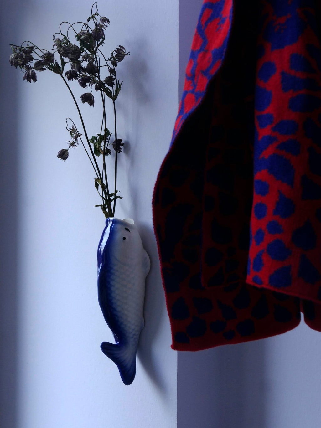 Detail von gestricktem Schal mit Blütenmuster in rot und indigo neben einer Keramik-Vase in Form eines Fisches die an der Wand hängt.