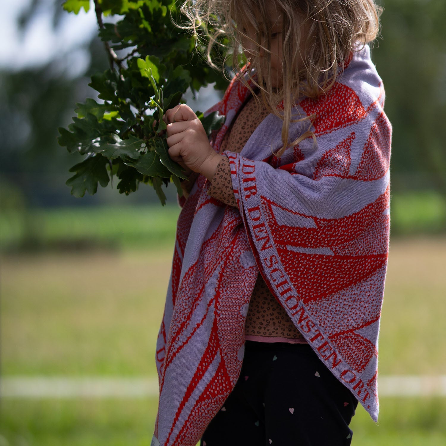 Kleinkind hat personalisierte lilimoon Babydecke gestrickt aus Baumwolle und Kaschmir in rot und flieder wärmend um die Schultern gelegt und erkundet neugierig die Natur.