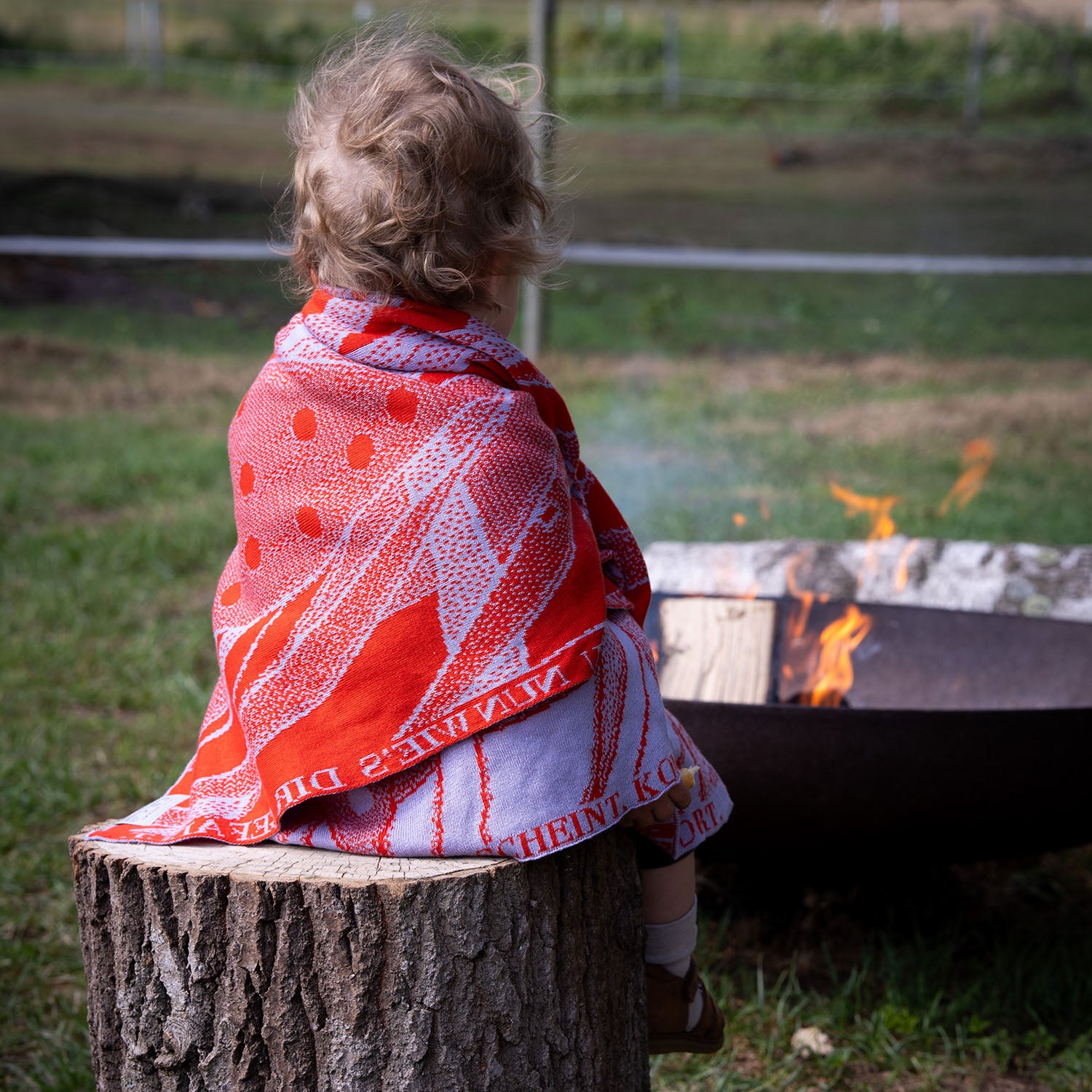 Kleinkind sitzt mit einer personalisierten lilimoon Babydecke gestrickt aus Baumwolle und Kaschmir in Rot und Flieder auf einem Baumstumpf vor einer Feuerstelle in der Natur.