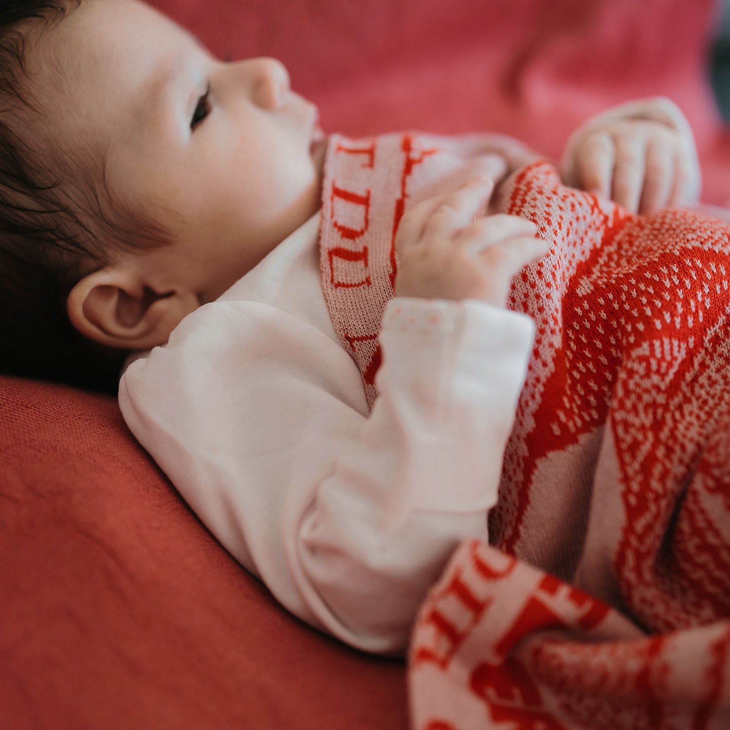 Ein Baby ist zugedeckt mit einer personalisierten lilimoon Babydecke gestrickt aus hochwertiger Baumwolle mit Kaschmir in rot und rosa.