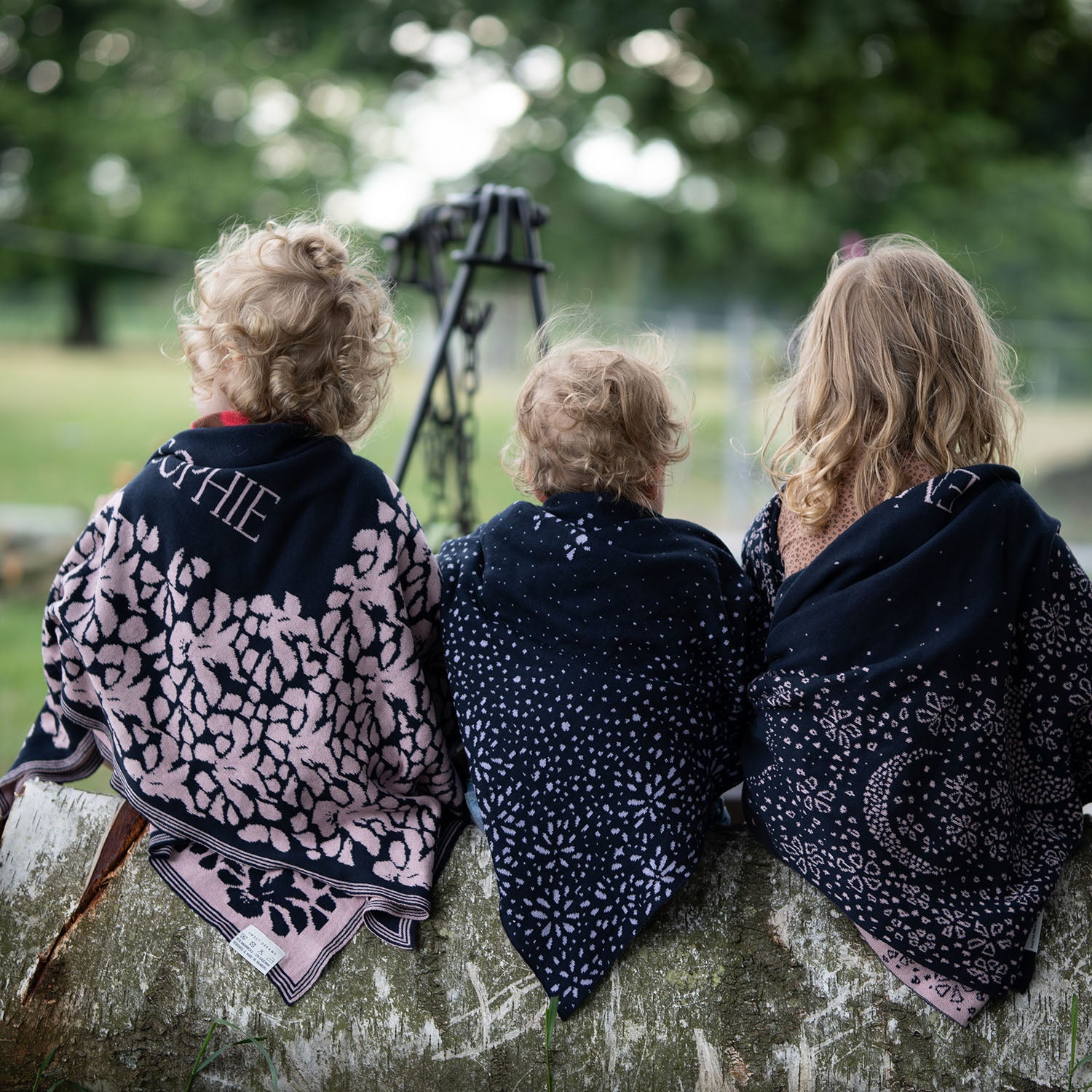 Drei Kleinkinder sitzen in der Natur auf einem Baumstamm und tragen gestrickte lilimoon Babydecken mit Sternen und Blumen wärmend über ihren Schultern.