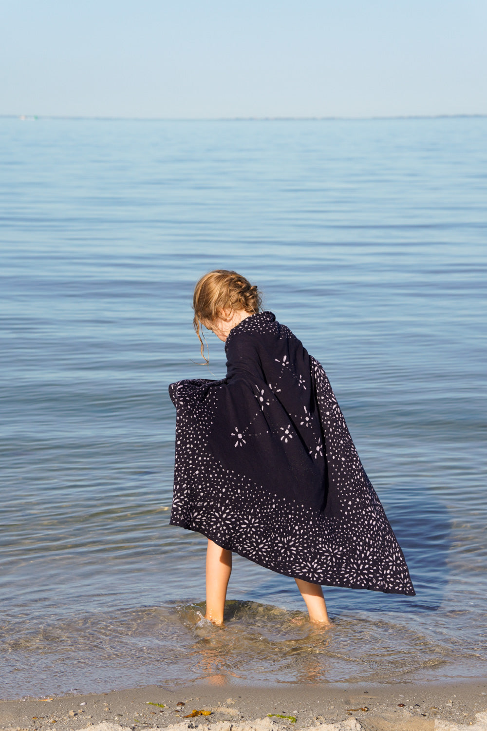 Kleinkind steht barfuss im Wattmeer der Nordsee und hat eine gestrickte lilimoon Babydecke mit Sternen und dem Sternzeichen Löwe als Umhang über die Schultern gelegt.