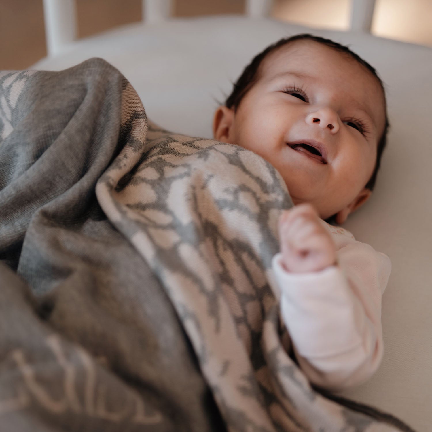 Ein fröhliches Baby liegt zugedeckt mit einer personalisierten lilimoon Babydecke in hellgrau und cremeweiss mit Blütenmuster im Kinderbett.