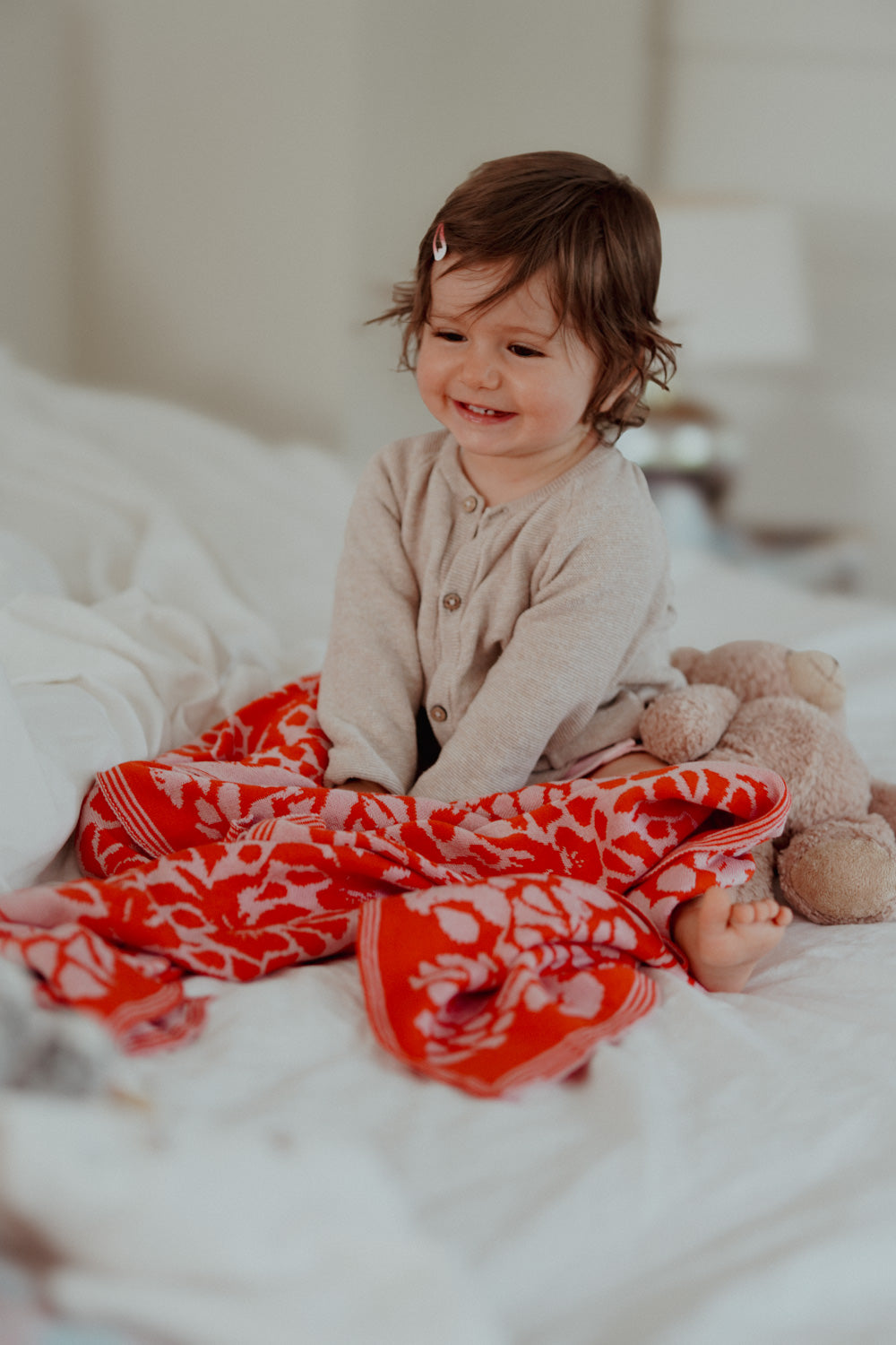 Ein fröhliches Kleinkind sitzt auf dem Bett und ist zugedeckt mit einer leuchtend rot und rosa farben lilimoon babydecke mit blütenmuster.