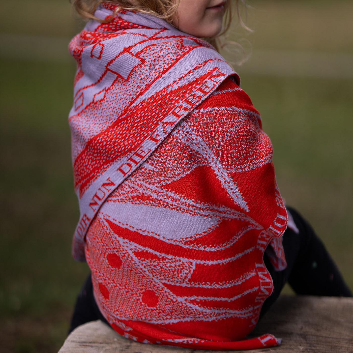 Kleinkind sitzt in der Natur umwickelt von einer personalisierten lilimoon Babydecke gestrickt aus Baumwolle mit Kaschmir in Flieder und Rot .